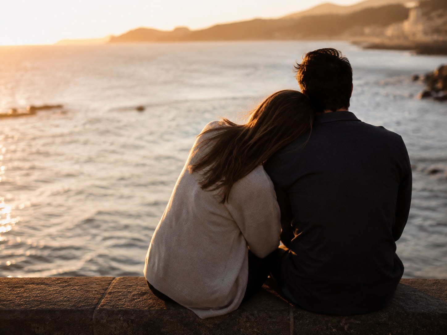 A couple sitting close on a stone sea wall, seen from behind, watching the water at sunset